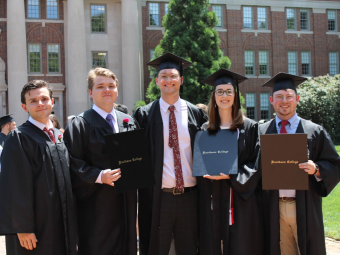 Five graduates in black academic caps and gowns pose outside holding Davidson College diploma covers in front of a brick campus building.