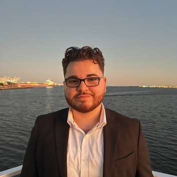 a young Middle Eastern man wearing glasses and a jacket in front of a body of water at sunset