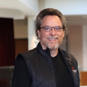 an older white man wearing a black shirt and a black vest with glasses