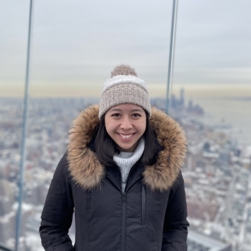 a young woman stands on the roof of a skyscraper in New York City, she wears a beanie and big coat and the sky appears to be grey and cloudy