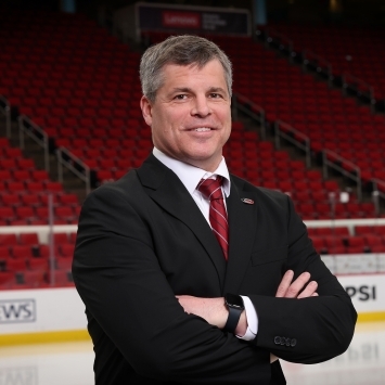 a middle aged white man wearing a suit and red tie in front of a hockey rink