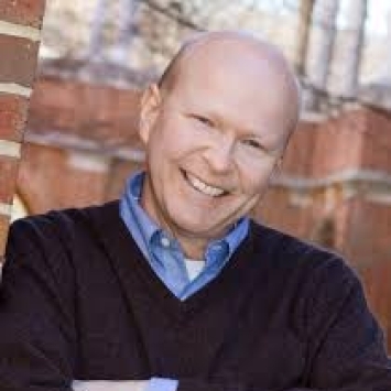 a white man wearing a collared shirt and sweater in front of a brick building
