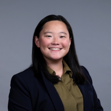 Smiling headshot of seated woman with black hair, green button down shirt, black blazer