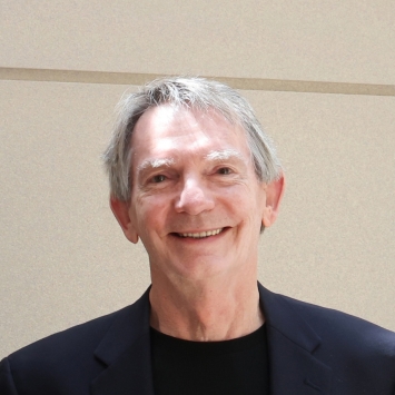 A headshot of a smiling older man with grey hair wearing a black shirt and a dark navy blazer against a neutral wall.