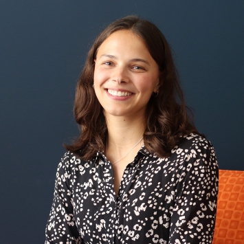 A smiling woman with shoulder-length wavy brown hair wearing a black and white patterned blouse, seated in front of a dark blue wall and an orange textured chair.