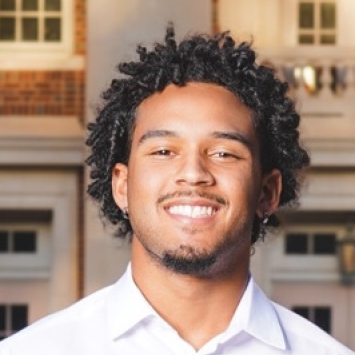 Headshot of a smiling young man with dark curly hair and a goatee, wearing a white collared shirt, in front of a blurred brick building with large windows.