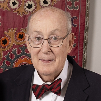 Portrait of an older man with glasses wearing a navy blazer, white shirt, and a red and black striped bowtie against a patterned tapestry background.