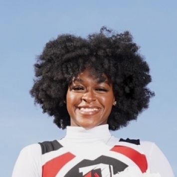 A close-up headshot of a smiling woman with voluminous dark hair wearing a white, black, and red athletic uniform against a clear blue sky.