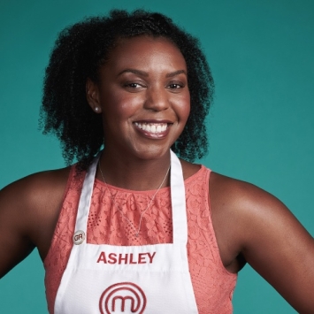 A smiling Black woman with curly hair wearing a white MasterChef apron with the name "ASHLEY" printed on it over a coral lace top, posed against a solid teal background.