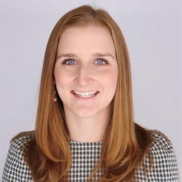 A smiling woman with long strawberry-blonde hair and blue eyes wearing a grey and white houndstoop-patterned top against a plain white background.
