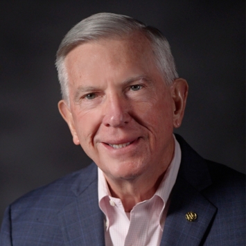 Portrait of Tom Ross wearing a blue windowpane patterned blazer and a light pink button-down shirt against a dark background.