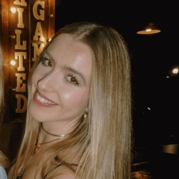 A smiling young woman with long, straight blonde hair and gold jewelry poses in a dimly lit indoor setting featuring a glowing sign in the background.