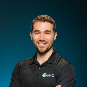 Professional headshot of a smiling man with short brown hair and a beard, wearing a black polo shirt featuring the "Lucid Bots" logo, standing with arms crossed against a teal gradient background.