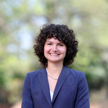 A smiling woman with dark curly hair and a pearl necklace sits outdoors wearing a navy blue blazer.