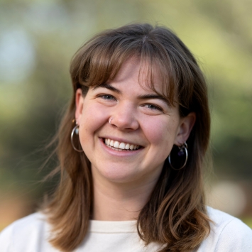 A close-up headshot of a smiling woman with light brown hair and bangs, wearing silver hoop earrings and a cream-colored top.