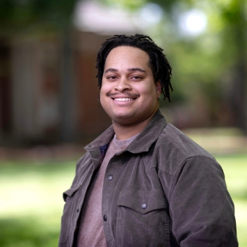 An outdoor portrait of a smiling man with a mustache and short dreadlocks, wearing a brown jacket over a lighter shirt, with a softly blurred background of trees and grass.