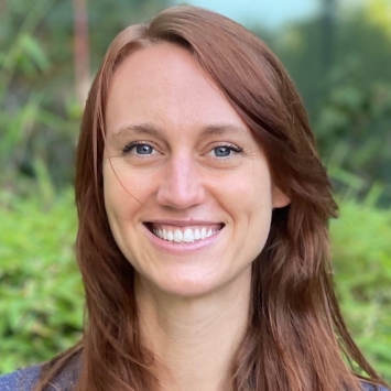 A close-up headshot of a smiling woman with long auburn hair and a blue blouse, posed against a blurred green foliage background.