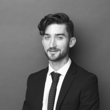 A black and white studio headshot of a man with styled dark hair and a light beard, wearing a dark suit jacket, white dress shirt, and a textured tie against a solid gray background.