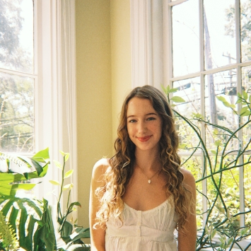 A young woman with long, curly blonde hair smiles gently while posing near a bright window flanked by large houseplants.