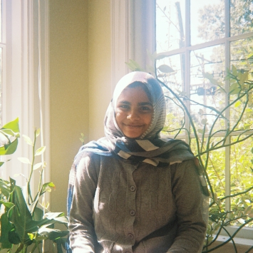 A smiling young woman wearing a patterned hijab and a grey button-up cardigan sits in a sunlit room filled with green plants.
