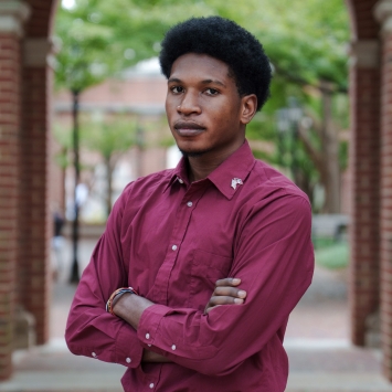 A young Black man with an afro stands with his arms crossed, wearing a maroon button-down shirt, framed by a brick archway on a college campus.
