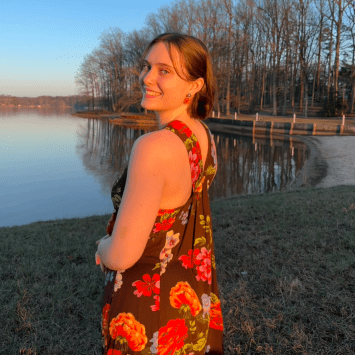 A young woman with short hair looks back smiling while wearing a floral-patterned dress in front of a quiet lake at sunset.