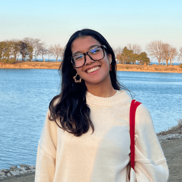 A smiling young woman wearing glasses and an oversized cream-colored sweater stands outdoors on a grassy slope next to a large body of water, with a tree-lined shore visible across the water under a clear blue sky.