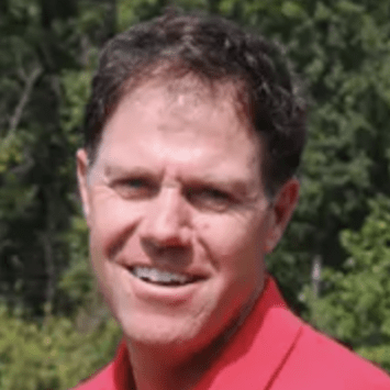 Close-up headshot of a smiling man with short brown hair, wearing a bright red polo shirt outdoors against a background of green trees.