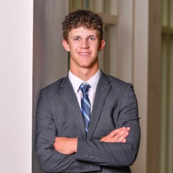 Headshot of a smiling young man with short curly brown hair, wearing a grey suit jacket, white dress shirt, and blue patterned tie, leaning against a white pillar with his arms crossed.