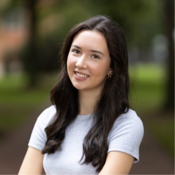 Headshot of a smiling young woman with long dark hair, wearing a light grey t-shirt, posed outdoors against a soft-focus green background.
