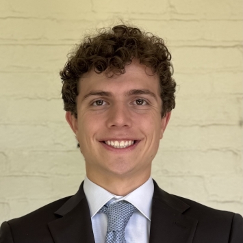 A young man with curly brown hair smiles directly at the camera while wearing a suit and blue tie
