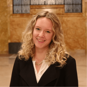 A woman with blonde curly hair smiles while wearing a black blazer over a white top and gold necklace