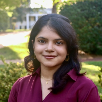 Mishti smiling outdoors in a deep plum-colored collared shirt with shoulder-length dark hair