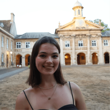 A smiling woman with shoulder-length brown hair wearing a thin black strap top stands in front of a historic stone building with a clock tower