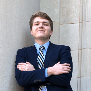 A smiling young man in a navy blazer, light blue dress shirt, and striped tie stands outdoors against a stone building wall with his arms crossed.