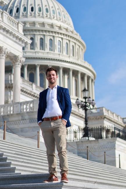 a young man wearing a jacket and collared shirt stands on the steps of the U.S. Capitol