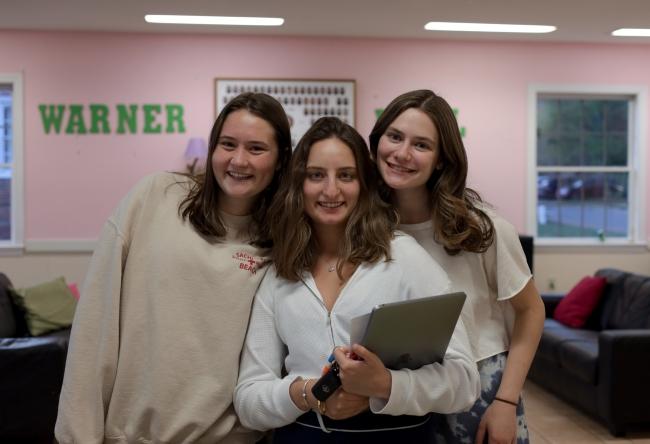 a group of three young women smiling in front of a pink wall that reads "Warner"