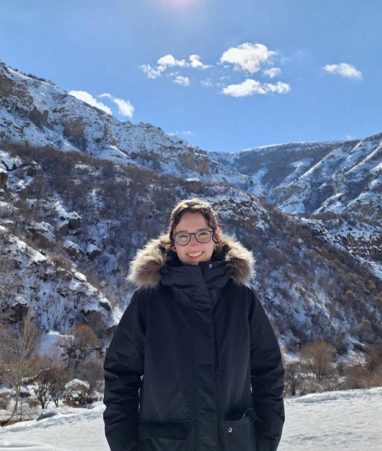 A white woman wears glasses and a big winter coat, stands smiling in front of mountains