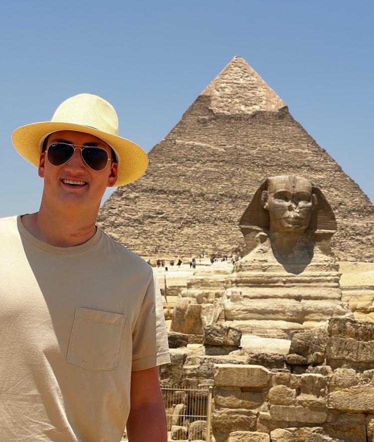 a young white male wears a straw hat while standing in front of an Egyptian pyramid and sphinx