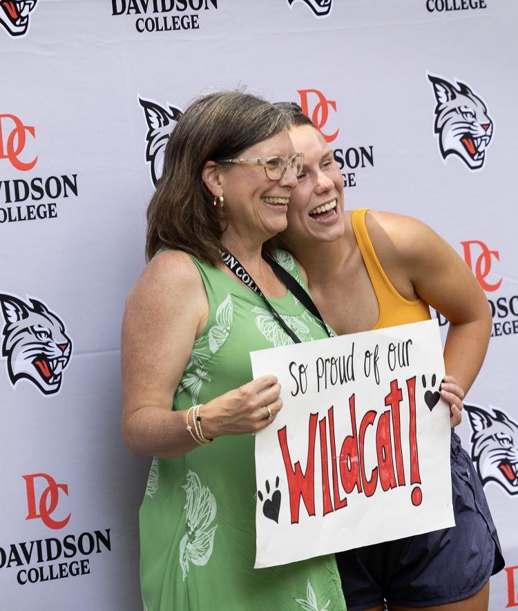 a mother and daughter stand together holding a sign that reads "so proud of our Wildcat!"