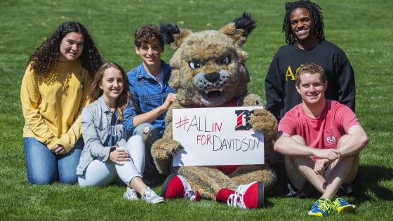 Lux and students sit on grass with sign reading #AllInForDavidson