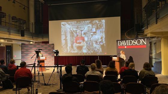students seated in the 900 room, looking toward the front at Tom Marshburn on a screen in a Davidson t-shirt in a spaceship with two in-person people behind podiums on both sides of the screen