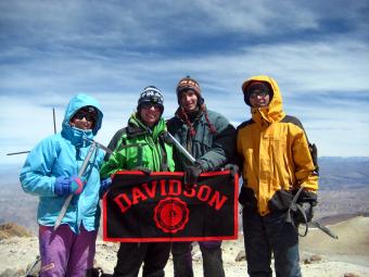 Four students in hiking gear stand atop a mountain in Peru holding a Davidson flag
