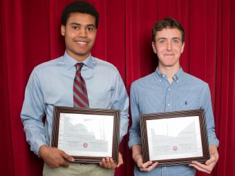 Two students stand in front of red curtain holding their economics awards