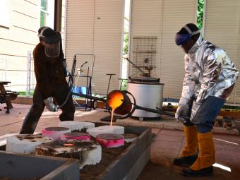Professor and student dressed in protective gear pour liquid bronze at the bronze foundry.
